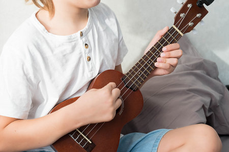 Young boy tuning ukulele at home. Blond haired boy sitting on couch playing acoustic guitar.