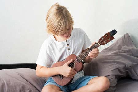 Young boy tuning ukulele at home. Blond haired boy sitting on couch playing acoustic guitar.の写真素材