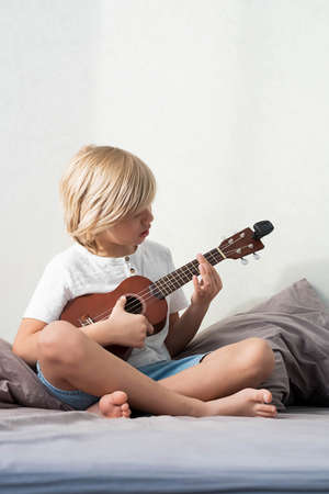 Young boy tuning ukulele at home. Blond haired boy sitting on couch playing acoustic guitar.の写真素材