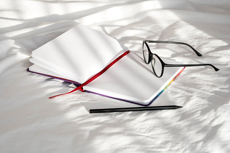 Blank notebook, pencil and eyeglasses rest on white bedding in sunlight with shadows. Stationary...
