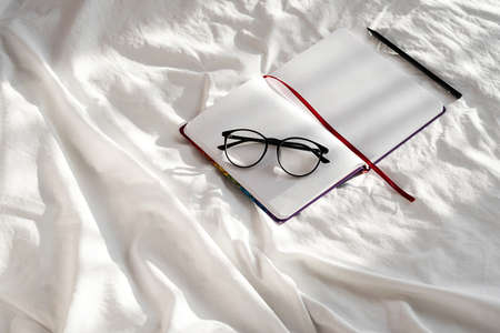 Diary, pencil and eyeglasses rest on white bedding in sunlight with shadows. Personal journal for notes. Stationary still life scene, morning at home.の写真素材