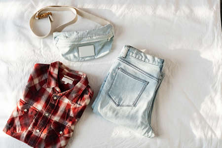 Blue jeans, waist bag and plaid shirt on white linen. Women's casual spring or summer outfit. Collage of everyday clothing, top view.の写真素材