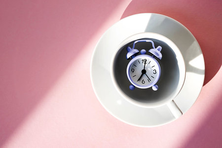 Violet vintage alarm clock in coffee cup on pastel pink background. Breakfast time concept, morning,...
