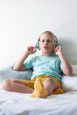Child wearing headphones listens to music. Cute boy listening to music in earphones seating on bed.の写真素材