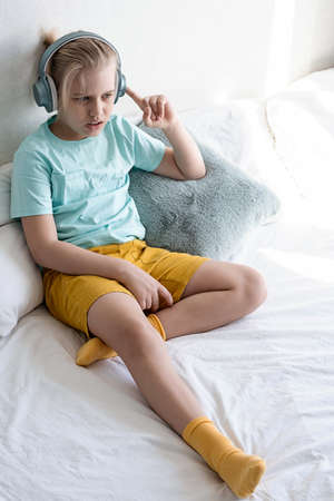 Cute boy listening to music in earphones seating on bed. Child wearing headphones listens to music.の写真素材