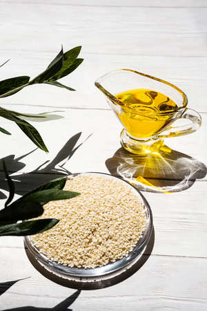 Sesame oil and heap of sesame seeds on white table. Still life of sesame oil and seeds in glass bowl, copy space.の写真素材