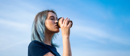 Blue haired woman drinks take away coffee against blue sky.の写真素材