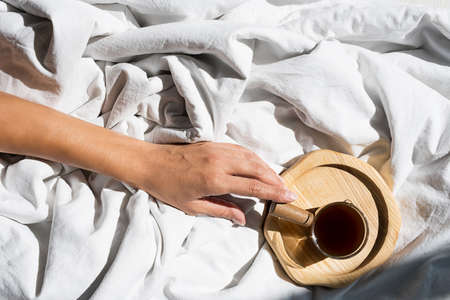 Females hand reaches for morning coffee cup in bed. Woman wakes up on white bedding in sunlight.の写真素材