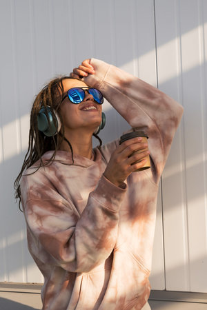 Dreadlock woman listening to music in headphones and drinking to go coffee, enjoying sunlight.