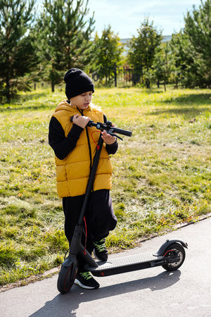 Boy rides an electric scooter in autumn park. Schoolboy using e-scooter at sunny day.