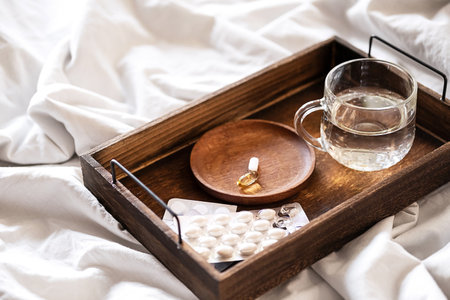 Wooden tray with pills, vitamins or supplements and water glass on the bed. Health care in the morning at home.の写真素材