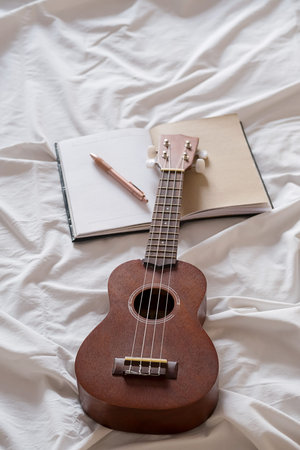 Ukulele guitar and open notebook on white bed. Cozy atmosphere for writing music.