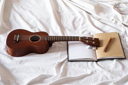 Ukulele guitar and open notebook on white bed. Cozy atmosphere for writing music.
