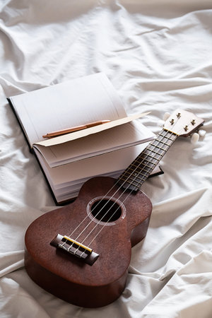 Ukulele guitar and open notebook on white bed. Cozy atmosphere for writing music.の写真素材