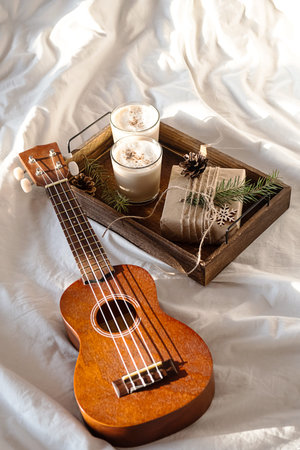 Christmas cozy morning at home. Gift box and eggnog on wooden tray near ukulele guitar on bed.
