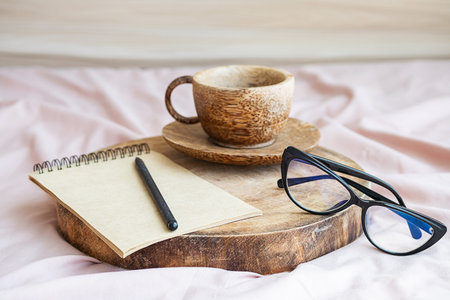 Morning coffee cup near blank notebook and goggles on wooden tray on sheet bedding.の写真素材