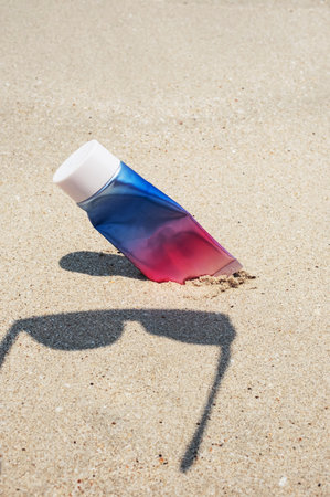 Used cosmetic sunscreen tube and sunglasses shadow on sandy beach.