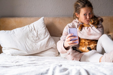 Woman taking selfie with cat on bed, making funny face. Cozy setting at home.の写真素材