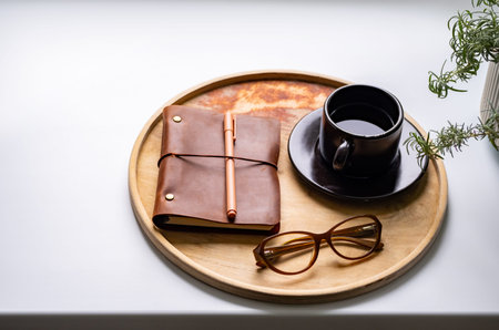 Cozy workspace with notebook, glasses and coffee cup on wooden tray.