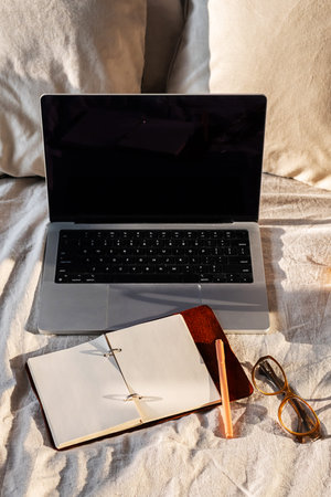 Open laptop, blank notebook and eyeglasses on cozy bed. Minimalist workspace vibe ideal for...