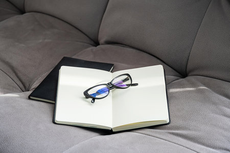 Open Notebook with Blank Pages and Pair of Glasses Rests on a Gray Couch. Cozy and Inviting Scene, Perfect for Studying or Writing.の写真素材
