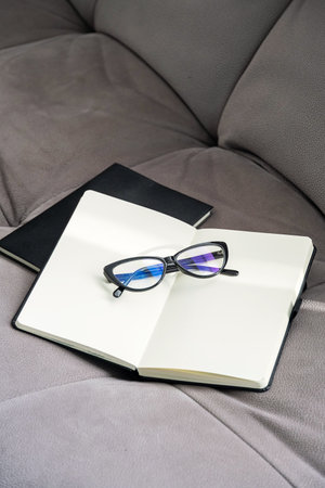 Pair of Stylish Black Glasses Rests on an Open Notebook Placed on Soft Gray Couch.の写真素材