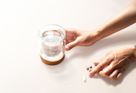 Hand Holding a Glass of Water Next to Several Pills on Light Surface. Healthcare and Wellness Concepts.の写真素材