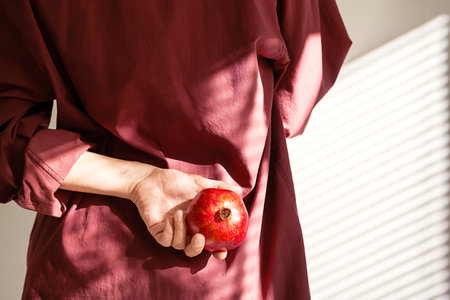 Woman Wearing Burgundy Shirt Holding Red Pomegranate Fruit Behind Back.の写真素材