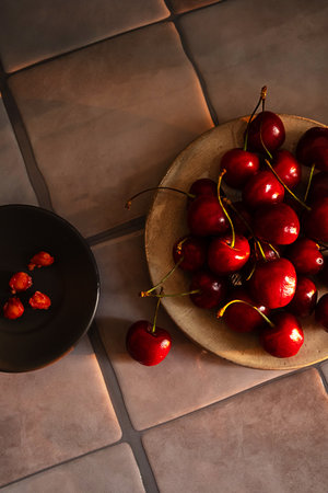 Fresh Juicy Red Cherries on Beige Ceramic Plate Next to Bowl with Cherry Pits on a Tile Surface.の写真素材