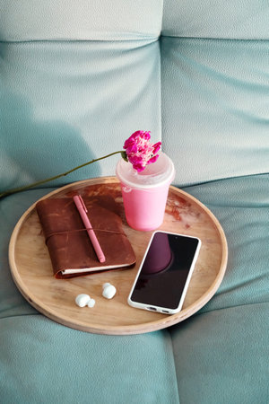 Overhead Shot of Wooden Tray with Notebook, Pen, Phone, Earbuds, and Pink Drink Cup with Flower on Sofa. Home Comfort Concept.