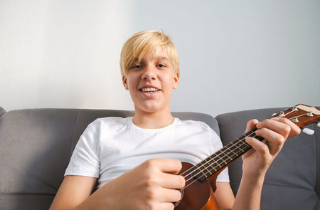 Caucasian Boy Playing Ukulele while Sitting on Sofa, Looking into Camera. Young Musician Learning to...