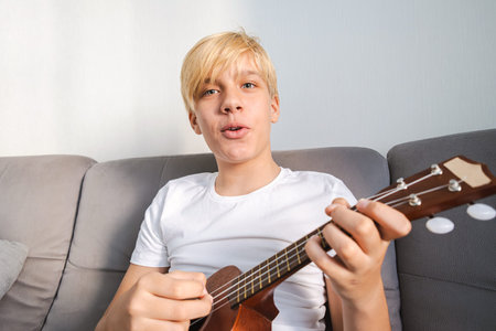 Caucasian Teen Boy Playing Ukulele and Singing a Song at Home. Music Education and Hobby Concepts.