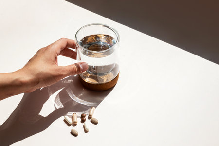 Person Reaching for Water Glass next to Scattered Vitamin Pills. Daily Supplement Intake for Health and Wellness.の写真素材