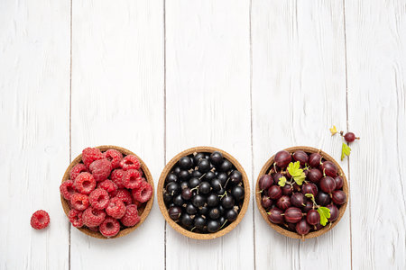 Three bowls filled with fresh raspberries, black currants and gooseberries on wooden background.の写真素材