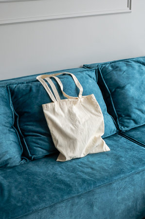 Reusable white fabric tote bag on a blue sofa. Empty textile shopper for eco-friendly shopping....