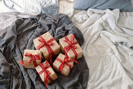Christmas gifts wrapped in kraft paper with red plaid ribbon on bed with linen duvet cover. Holiday surprise concept.の写真素材