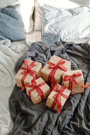 Wrapped brown paper gift boxes with red plaid ribbon on gray rumpled bed. Christmas morning present...