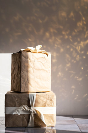 Stacked gift boxes wrapped in brown kraft paper with white ribbon, on reflective surface with...