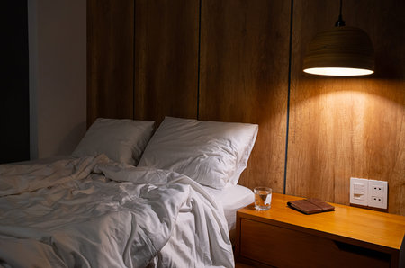 Unmade bed with white linen and pillows in hotel room with glass of water, book on nightstand at night, cozy atmosphere.の写真素材