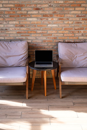 Laptop with blank screen on small coffee table between two armchairs, against brick wall. Remote work setup for modern lifestyle.の写真素材
