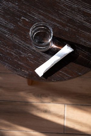 Glass of water and white powder sachet on dark wooden table in natural light. Nutrition supplement...