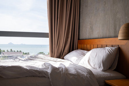 Cozy bed with white sheets and pillow in sunny room by large window overlooking the sea, perfect tranquil bedroom scene for relaxation.の写真素材
