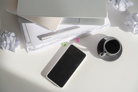 Laptop, stack of paper, crumpled paper, and smartphone on desk for business work. Freelance and...