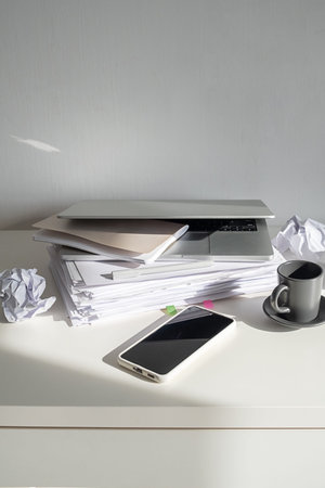 Laptop on stack of paper with coffee cup and smartphone on desk. Workplace clutter with pile of...