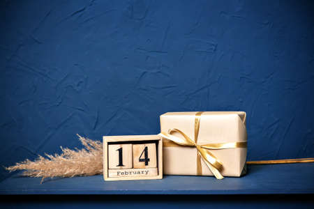Gift for Valentines Day. The box is wrapped with kraft paper. Golden bow. Fluffy dry grass feather grass. Blue table or chest of drawers. Textured plaster on the wall in dark blueの写真素材