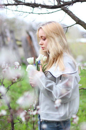 A beautiful young girl stands among the flowering trees. White flowers. Spring. A girl with white hair, in a transparent jacket, stands near a blossoming tree with white tulips. Spring moodの写真素材