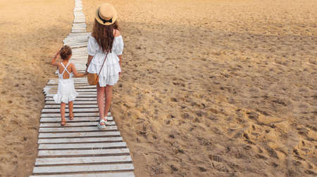 two girls in white dresses are walking along the beach path on the yellow sand. back viewの写真素材