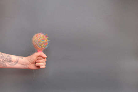 Hair loss in comb. female hand with a tattoo holds a pink comb with white hair on a gray background. Hair health conceptの写真素材