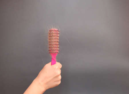 Hair loss in comb. female hand with a tattoo holds a pink comb with white hair on a gray background. Hair health conceptの写真素材