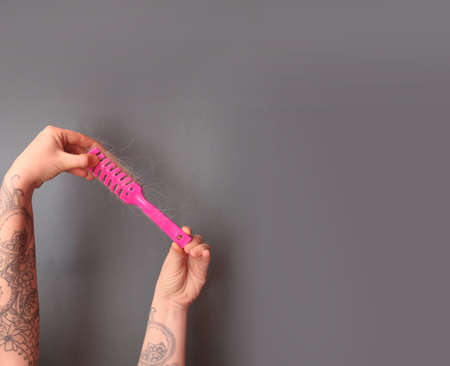 Hair loss in comb. female hand with a tattoo holds a pink comb with white hair on a gray background. Hair health conceptの写真素材